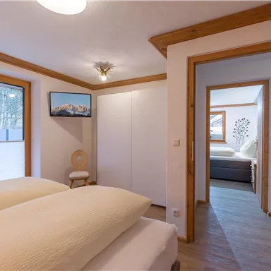 A modern bedroom with two beds and large windows. The room has light walls and cozy wooden details.