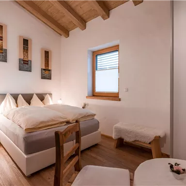 A cozy bedroom with a double bed and a light wooden ceiling. The walls are plain, and there is a small table and chairs.