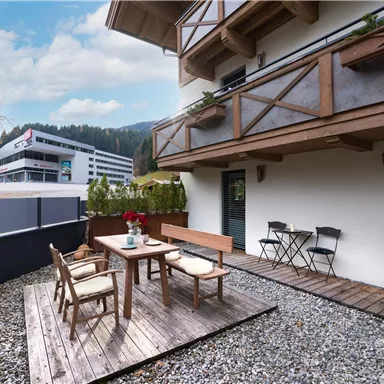 A cozy outdoor area with a wooden terrace, table, and chairs. In the background, mountains and modern buildings can be seen.