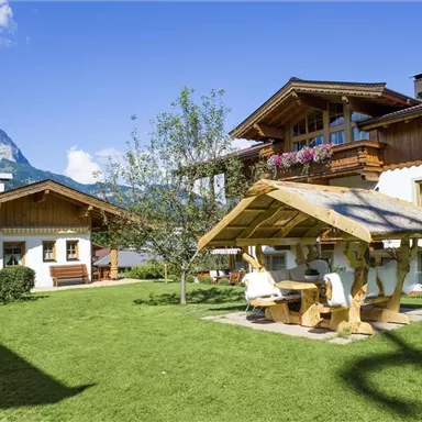 Ein schöner Garten mit zwei traditionellen Häusern und einem Holzpavillon. Im Hintergrund sind Berge und ein blauer Himmel zu sehen.