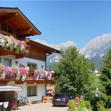 Ein charmantes alpenländisches Haus mit blühenden Balkonen. Im Hintergrund sind hohe Berge und ein klarer blauer Himmel zu sehen.