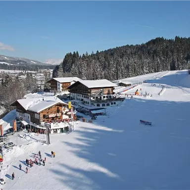 A snow-covered landscape with cozy cabins and people skiing. The clear blue sky and the wooded mountains create an idyllic winter atmosphere.