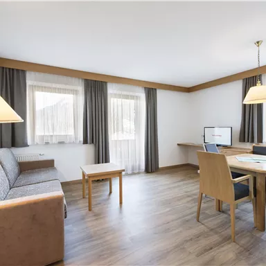 A cozy living room with a sofa, a table, and a television. Large windows provide plenty of natural light.