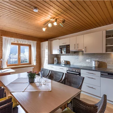 Eine moderne Küche mit Holzdecke und weißen Schränken. Ein Esstisch mit Stühlen steht im Vordergrund, begleitet von einer dekorativen Pflanze.