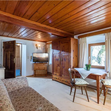 Ein gemütliches Wohnzimmer mit Holzdecke und einer Sitzecke. Es gibt einen Tisch, Stühle und einen Fernseher, sowie viel Tageslicht durch das Fenster.