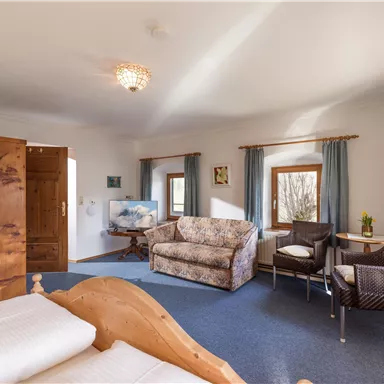 Ein gemütliches Zimmer mit einem Doppelbett und einem bequemen Sofa. Es gibt Fenster mit Vorhängen und einen kleinen Tisch mit Stühlen.