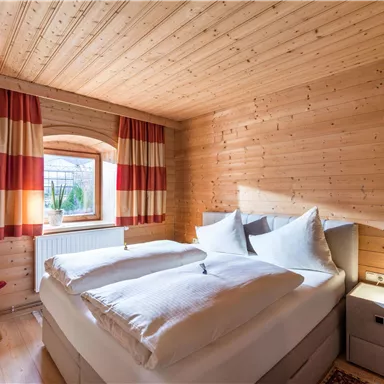 Ein gemütliches Schlafzimmer mit Holzverkleidung und großen Fenstern. Das Bett ist mit weißen Kissen und Decken ausgestattet, und die Vorhänge haben ein rot-weißes Streifenmuster.
