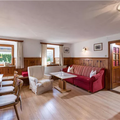 Ein gemütlicher Wohnraum mit Holzverkleidung und großen Fenstern. Ein rotes Sofa und ein Esstisch mit Stühlen laden zum Entspannen ein.