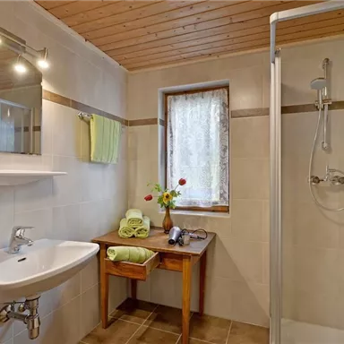 A modern bathroom with a shower, a sink, and a wooden shelf. There are fresh towels and a small floral decoration on the table.