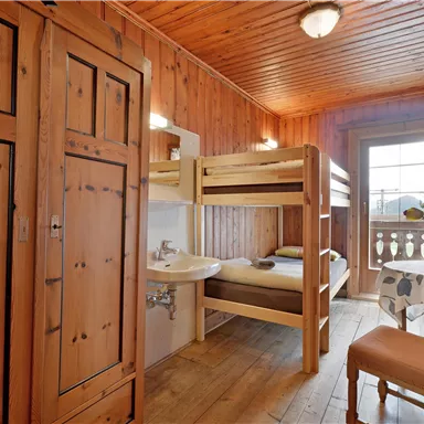 A cozy wooden room with a bunk bed and a desk. The window offers a view of the balcony and the surrounding landscape.