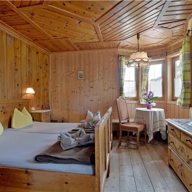 A cozy wooden room with two beds and yellow cushions. A table with chairs and a blooming vase completes the warm atmosphere.