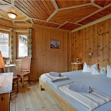 A cozy room with wooden paneling and a large bed. A table with a chair and gentle light create an inviting atmosphere.