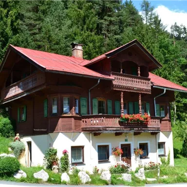A traditional wooden house with a red roof and blooming flowers. It stands amidst green trees and a picturesque landscape.