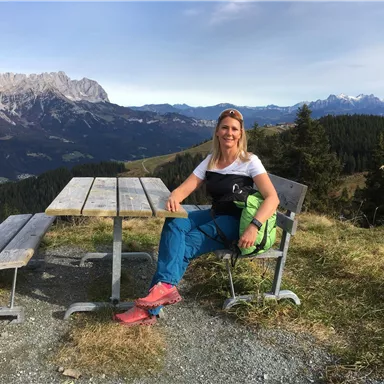 Eine Frau sitzt an einem Tisch mit Blick auf die Berge. Die Landschaft ist von Bäumen und Alpen geprägt.