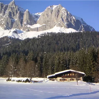 A snow-covered valley with a charming mountain cabin in the foreground. In the background, majestic mountains rise under a clear blue sky.