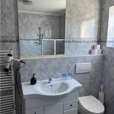 A modern bathroom with a white sink line and a large mirror. The walls are tiled in light colors, and there is a shower as well as hygiene products.