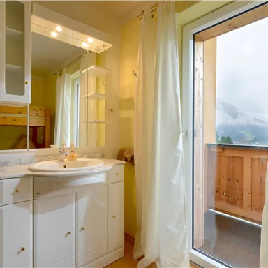 A bright bathroom with a sink and a large window. Through the window, a balcony with a mountain view can be seen.