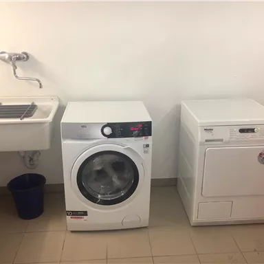 Ein Waschraum mit einer modernen Waschmaschine und einem Wäschetrockner. Neben den Geräten steht ein Waschbecken und ein blauer Eimer.