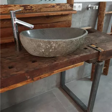 A modern washbasin made of wood with an elegant stone bowl. In the background, gray tiles and rustic wood details are visible.