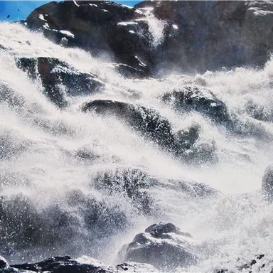 Stark tosende Wellen, die gegen große Felsen schlagen. Spritzendes Wasser und ein dramatischer Himmel im Hintergrund.