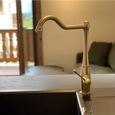 Ein eleganter Wasserhahn aus Messing steht über einer modernen Spüle. Im Hintergrund ist ein gemütlicher Wohnbereich mit einem Sofa zu sehen.