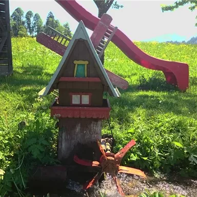 A colorful playhouse with a water wheel stands in a green garden. In the background, you can see a slide and plenty of blooming meadow.