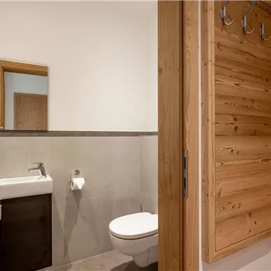 A modern bathroom with a toilet, a sink, and wooden details. Towels are hanging on the wall and there is a wooden dressing area.