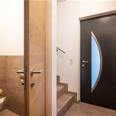 Ein moderner Eingangsbereich mit einer türförmigen schwarzen Tür und einer hellen Treppe. Rechts ist eine kleine Toilette mit einem Handtuch sichtbar.