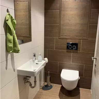 A modern toilet with brown tiles and a small sink. A green towel hangs on the wall.