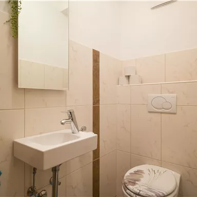 Eine kleine, helle Toilette mit weißen Fliesen und einem Waschbecken. An der Wand hängt ein Handtuch in Türkis.