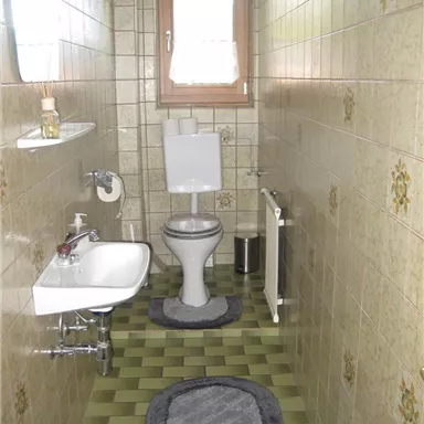 A small bathroom with a toilet, a sink, and a window. The walls are tiled, and the floor has a green pattern.