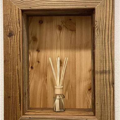 Ein dekorativer Holzrahmen mit einer hellen Holzplatte im Hintergrund. In der Mitte steht ein Flakon mit Duftsticks.