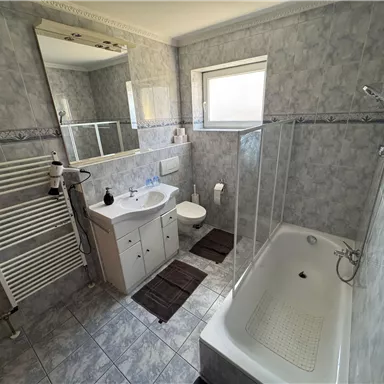A modern bathroom with shower and bathtub.  
Tiles in gray and a large mirror create a bright atmosphere.