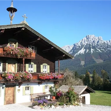 Ein traditionelles Holzhaus mit blühenden Blumen auf den Balkonen. Im Hintergrund sind majestätische Berge zu sehen.