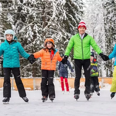 Eine fröhliche Gruppe von Kindern und einem Erwachsenen genießt das Eislaufen auf einer gefrorenen Fläche im Schnee. Alle tragen bunte Winterjacken und Helme.