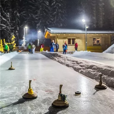 Eine Winterlandschaft mit einem beleuchteten Eisfeld und Menschen, die sich im Hintergrund aufhalten. Im Vordergrund sind mehrere Holzscheiben zum Spiel zu sehen.