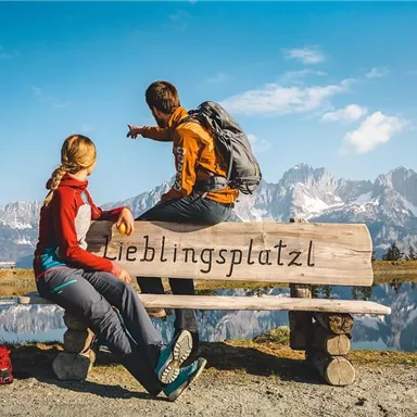A couple sits on a bench with the inscription "Favorite Place" and looks out at the picturesque mountain landscape. The sky is clear and the surroundings are idyllic.