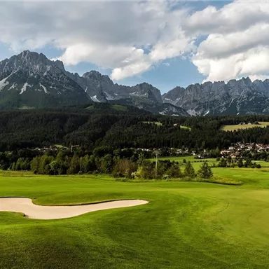 Eine grüne Golfplatzlandschaft umgeben von majestätischen Bergen. Der Himmel ist teilweise bewölkt, und der Ort strahlt Ruhe aus.