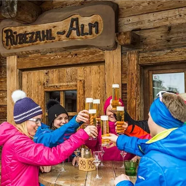 Eine Gruppe von Menschen hebt ihre Biergläser vor einer Berghütte. Sie tragen bunte Winterkleidung und feiern gemeinsam.