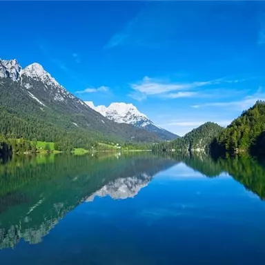 Ein klarer See umgeben von hohen Bergen und üppigem Grün. Der Himmel ist blau und strahlend, und die Landschaft spiegelt sich im Wasser.
