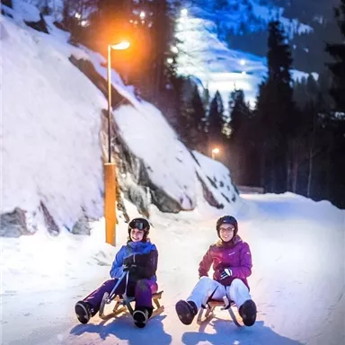 Zwei Personen sitzen auf Rodeln und genießen die nächtliche Winterlandschaft. Die verschneite Straße wird von Laternen beleuchtet, während im Hintergrund die Berge sichtbar sind.