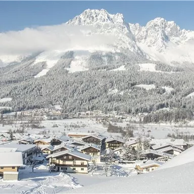 A snow-covered landscape with a picturesque village and high mountains in the background. The sky is clear, and there is a tranquil winter atmosphere in the air.