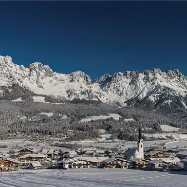 Ein malerisches Dorf in einer schneebedeckten Landschaft, umgeben von majestätischen Bergen. Der klare blaue Himmel vervollständigt die winterliche Szene.