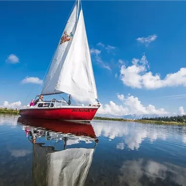 Ein Segelboot mit weißem Segel gleitet über einen ruhigen See. Der Himmel ist blau mit einigen Wolken und spiegelt sich im Wasser.
