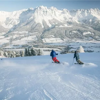 Zwei Skifahrer fahren auf einer verschneiten Piste mit beeindruckenden Bergen im Hintergrund. Der Himmel ist klar und die Landschaft ist von Schnee bedeckt.