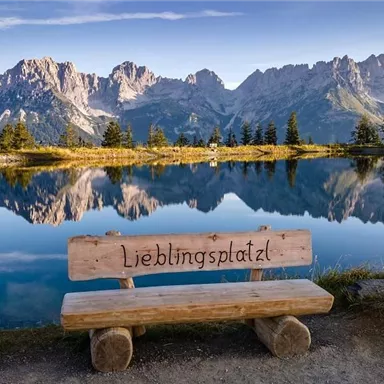 Eine wunderschöne Landschaft mit Bergen im Hintergrund und einem ruhigen See. Eine Holzbank mit der Aufschrift „Lieblingsplatz“ lädt zum Verweilen ein.