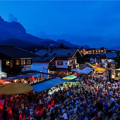 Eine lebhafte Menschenmenge versammelt sich an einem Abendmarkt. Im Hintergrund sind die Berge und beleuchtete Stände sichtbar.
