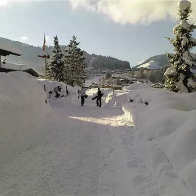 A snow-covered street with high snowbanks on both sides. People are walking in the wintry landscape on a sunny day.