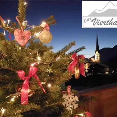 A decorated Christmas tree with lights and ornaments at night. In the background, there is a church with a tall tower and a clear night sky.
