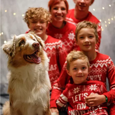 A cheerful family poses together with a dog in front of a festive backdrop. Everyone is wearing Christmas sweaters and smiling.